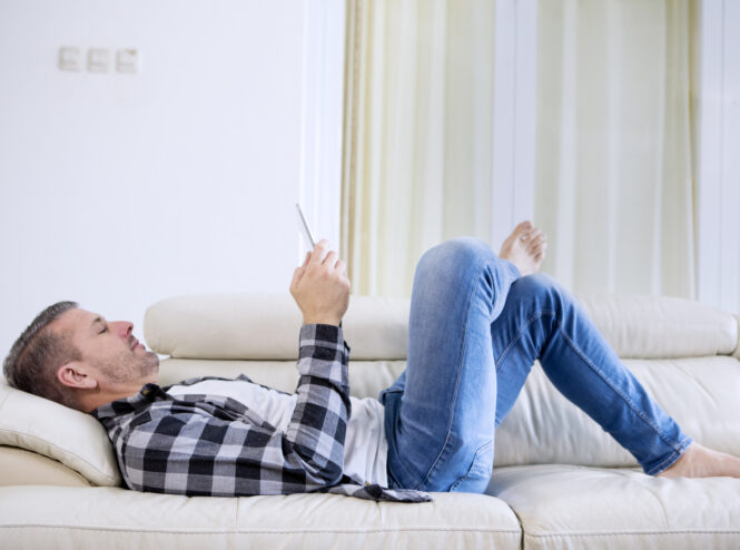 Middle aged caucasian man relaxing at home lying on sofa looking at digital tablet laughing